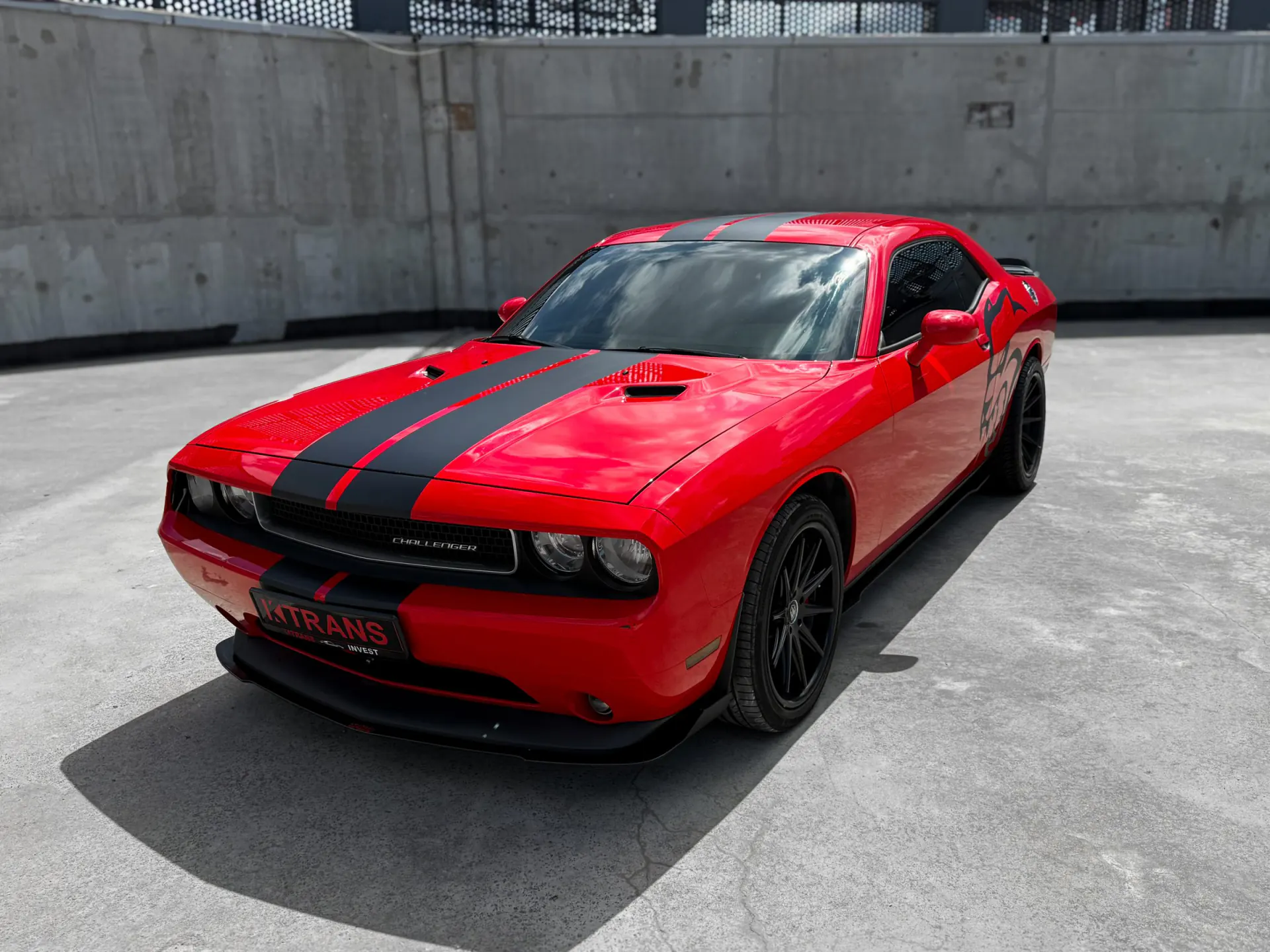 Червоний Dodge Challenger 2013 з чорними смугами й табличкою KTRANS на бетонному даху паркінгу